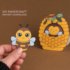Könnte beinhalten: Ein Bild eines Papierbastelprojekts mit einer Cartoon-Biene und einem Bienenstock. Die Biene ist gelb und braun mit blauen Flügeln, gehalten von einer Hand. Der Bienenstock ist gelb mit einem Wabenmuster und einer Biene im Inneren. Text: "DIY Papercraft Instant Download."