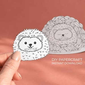 Könnte beinhalten: Ein weißer Papier-Igel mit einem lächelnden Gesicht wird von einer Hand gehalten, neben einer größeren Igelform aus Blättern. Der Text "DIY PAPERCRAFT INSTANT DOWNLOAD" ist sichtbar. Der Hintergrund ist einfarbig pfirsichfarben.