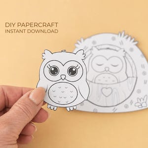 Könnte beinhalten: Ein schwarz-weißes DIY-Papierbastel-Eulen-Ausschnitt, der von einer Hand gehalten wird, mit einem zweiten Eulendesign im Hintergrund. Der Text "DIY PAPERCRAFT INSTANT DOWNLOAD" steht oben. Die Eule hat große Augen und einen herzförmigen Bauch.