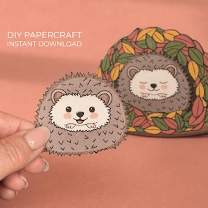 Könnte beinhalten: DIY-Papierbastelarbeit mit einem lächelnden Igel mit grauem Körper und weißem Gesicht, gehalten von einer Hand. Ein zweiter Igel ruht in einem laubbedeckten Bau. Der Text "DIY PAPER CRAFT INSTANT DOWNLOAD" ist sichtbar.