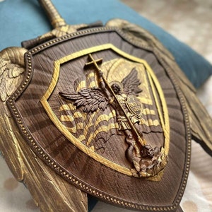 May include: A wooden shield with a gold-painted angel holding a sword and a cross. The shield is decorated with gold-painted wings and a gold-painted border. The shield is mounted on a wall.