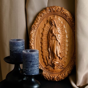 May include: Wooden oval plaque with a carved image of a religious figure, surrounded by an ornate floral border. Two dark blue pillar candles sit in black metal candle holders. The background includes beige fabric.