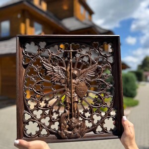 May include: A square dark brown wooden wall plaque featuring an intricate carved relief of Saint Michael the Archangel defeating a dragon. The central figure is framed by a detailed gothic-style openwork circular pattern with the text Quis Ut Deus.