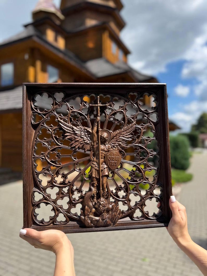 May include: A square dark brown wooden wall plaque featuring an intricate carved relief of Saint Michael the Archangel defeating a dragon. The central figure is framed by a detailed gothic-style openwork circular pattern with the text Quis Ut Deus.