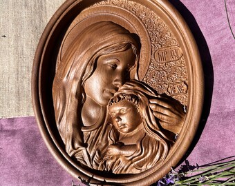 May include: A wooden carving of the Virgin Mary holding the baby Jesus. The carving is in a circular frame and has a detailed, ornate design. The carving is brown and has a natural wood finish.
