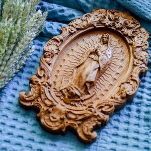 May include: Ornate, wooden religious plaque featuring a carved image of a religious figure within an oval frame. The frame has detailed scrollwork and is set against a blue textured background. Wheat stalks are visible in the upper left corner.