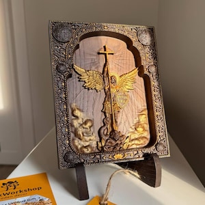 May include: A wooden relief sculpture of an angel with golden wings, holding a cross and shield, set within an ornate, dark brown frame. The artwork is displayed on a wooden stand, with a tag from "Wooden Decor" visible.