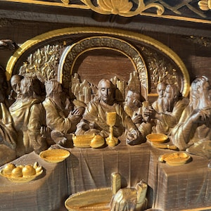 May include: A wooden relief sculpture depicting the Last Supper. The figures are carved in a realistic style and are painted in shades of brown and gold. The scene is set in a room with a table and chairs. The figures are seated around the table, and Jesus is in the center.