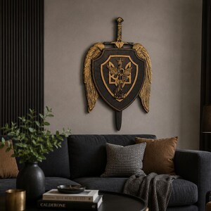 May include: A wall-mounted decorative piece featuring a black shield with gold-colored wings and a central gold trident emblem. A sword is positioned vertically behind the shield, with the hilt extending above and the blade tip visible below.
