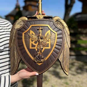 May include: A dark brown wooden shield featuring a central gold-toned relief of Archangel Michael standing over a serpent, with the Ukrainian trident symbol behind him. The shield is adorned with large gold-toned wings and a sword handle at the top.