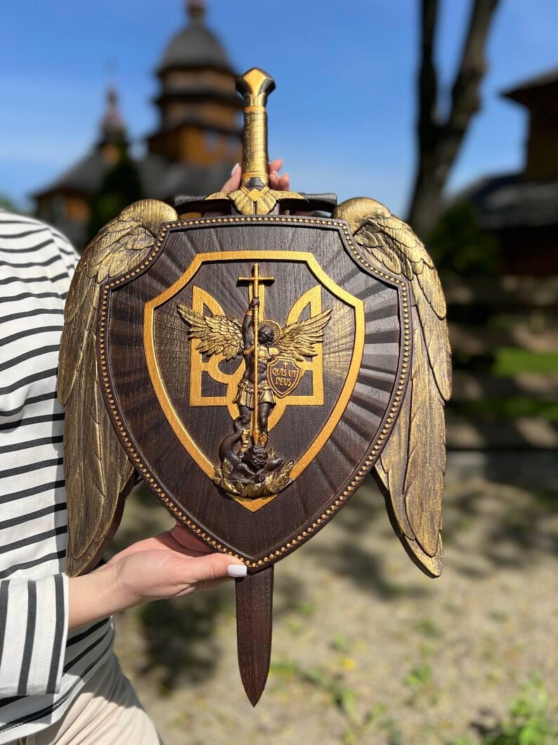 May include: A dark brown wooden shield featuring a central gold-toned relief of Archangel Michael standing over a serpent, with the Ukrainian trident symbol behind him. The shield is adorned with large gold-toned wings and a sword handle at the top.