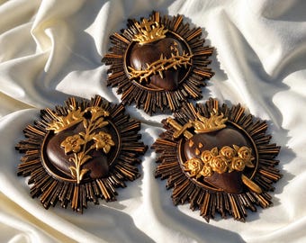 May include: Three circular wall plaques featuring dark brown heart-shaped centers with gold-painted accents. Each plaque has a radiating sunburst border. The designs include a crown of thorns, a floral stem, and roses with a sword.