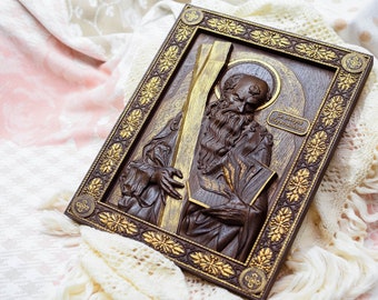 Puede incluir: Un icono rectangular de madera con un relieve tallado de un santo barbudo sosteniendo una cruz y un libro. El icono es principalmente marrón con detalles dorados en el marco y la cruz. El santo está representado con una aureola. El texto del icono está en un idioma extranjero.