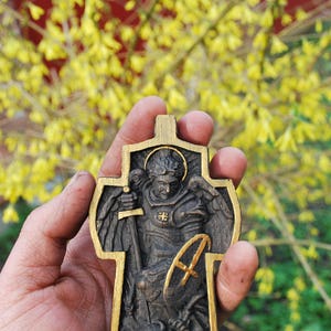 May include: A wooden cross pendant with a detailed relief carving of a saint holding a sword and shield. The cross has a gold-colored border and is held against a backdrop of yellow flowers.