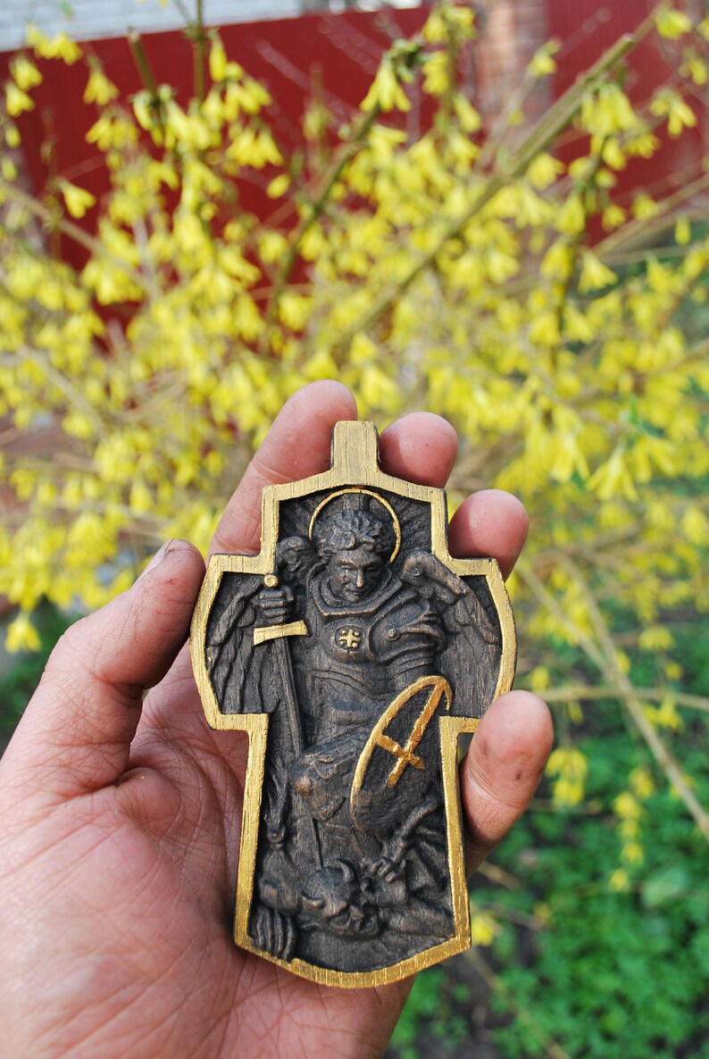 May include: A wooden cross pendant with a detailed relief carving of a saint holding a sword and shield. The cross has a gold-colored border and is held against a backdrop of yellow flowers.