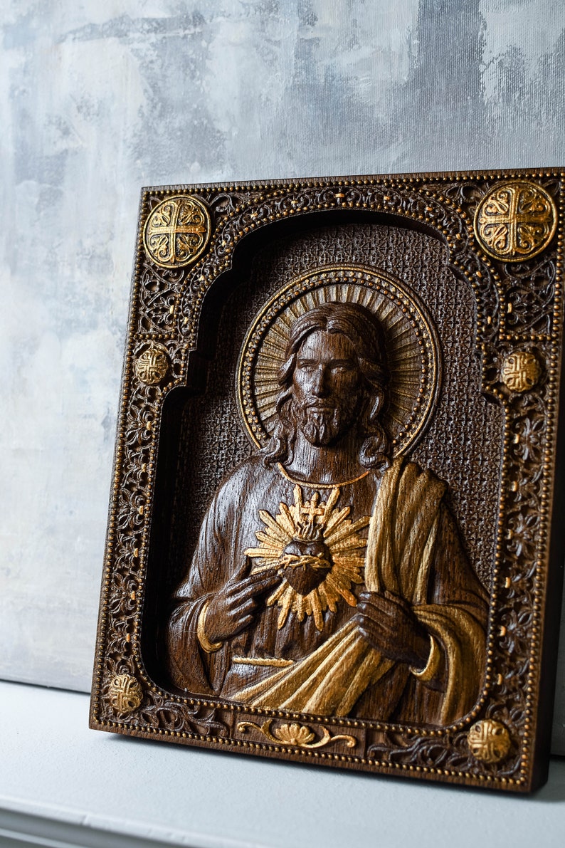 K&ouml;nnte beinhalten: Eine rechteckige Wandtafel aus Holz mit einem geschnitzten Relief des Heiligsten Herzens Jesu. Das dunkelbraune Holz ist mit goldener Farbe an der Figur, dem Herzen und dem verzierten Rand akzentuiert, der vier kreisf&ouml;rmige Kreuzmotive enth&auml;lt.