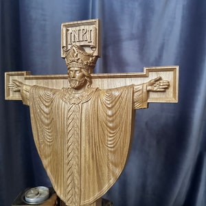 Może przedstawiać: Drewniana rzeźba krucyfiksu przedstawiająca ukoronowaną postać w szatach. Krzyż ma wyciągnięte ramiona, a nad głową widnieje napis "INRI". Rzeźba wykonana jest z jasnego drewna.