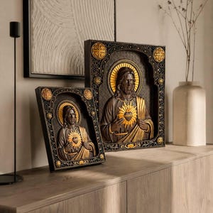 K&ouml;nnte beinhalten: Zwei rechteckige Wandtafeln mit einem Relief des Heiligsten Herzens Jesu. Die Figuren sind in Dunkelbraun und Gold gehalten und in aufwendige schwarze Rahmen mit goldenen kreisf&ouml;rmigen Akzenten an den Ecken und Seiten eingefasst.