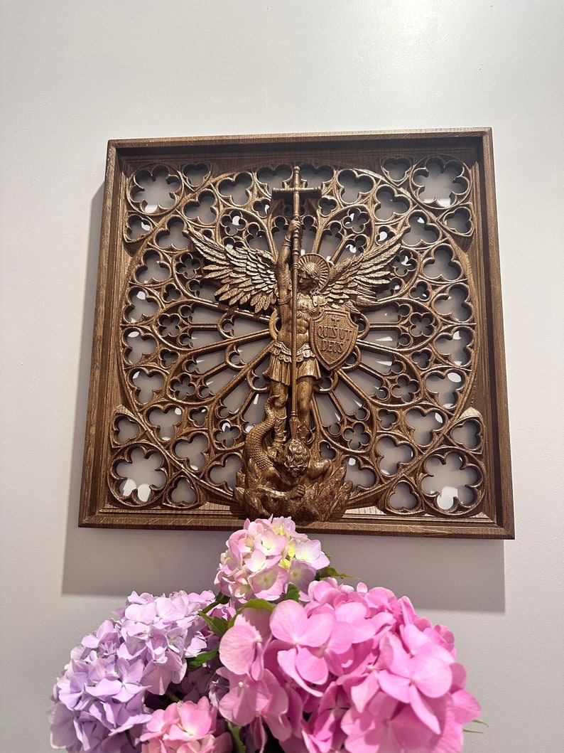 May include: A square brown wooden wall plaque featuring an intricate openwork rose window design. In the center, a relief carving depicts an angel with wings holding a long staff and a shield inscribed with the Latin text Quis Ut Deus.
