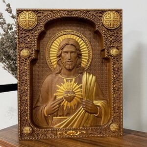 K&ouml;nnte beinhalten: Eine rechteckige Wandtafel aus Holz mit einem Relief des Heiligsten Herzens Jesu. Die Figur ist mittig mit einem goldenen Heiligenschein und Herzen dargestellt, umgeben von einem verzierten, tief geschnitzten Rahmen mit vier goldenen Akzenten.
