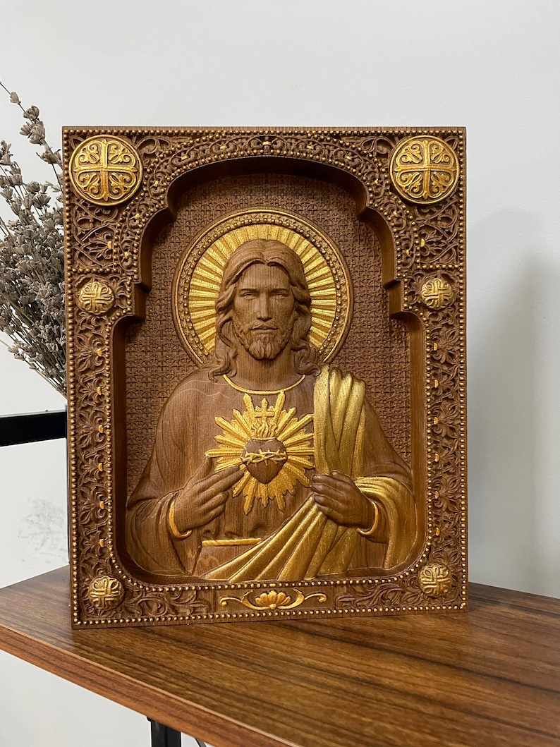 K&ouml;nnte beinhalten: Eine rechteckige Wandtafel aus Holz mit einem Relief des Heiligsten Herzens Jesu. Die Figur ist mittig mit einem goldenen Heiligenschein und Herzen dargestellt, umgeben von einem verzierten, tief geschnitzten Rahmen mit vier goldenen Akzenten.