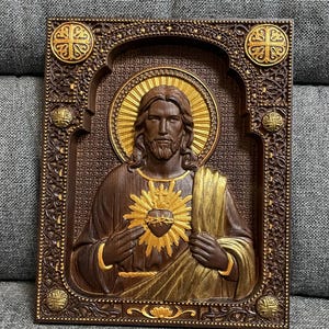 K&ouml;nnte beinhalten: Eine rechteckige Wandtafel aus Holz mit einem geschnitzten Relief des Heiligsten Herzens Jesu. Die Figur ist mit langem Haar, Bart und einem goldenen Heiligenschein dargestellt. Der Rahmen ist dunkelbraun mit floralen Schnitzereien.