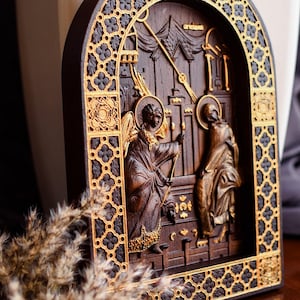 May include: A carved wooden icon depicting a religious scene with two figures. The icon is framed with a decorative border of gold and black. The icon is set against a background of a wooden surface.