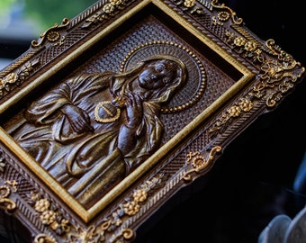 Puede incluir: Una talla de madera enmarcada de una mujer con halo, vestida con una túnica y sosteniendo un corazón. El marco es ornamentado con detalles dorados.