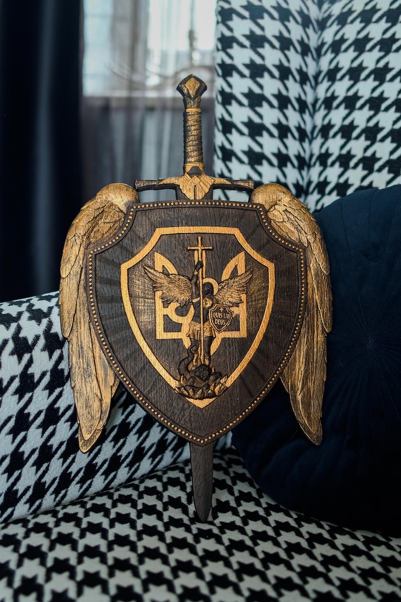 May include: A dark brown wooden shield featuring a central gold-toned relief of an angel with wings and a sword, overlaid on a trident symbol. The shield is flanked by two large gold-toned wings and topped with a sword hilt. The text "QUIS UT DEUS" is visible on a small shield held by the angel.