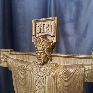 Może przedstawiać: Drewniany krucyfiks z wyrzeźbioną postacią Jezusa z wyciągniętymi ramionami. Postać nosi koronę i szatę. Na g&oacute;rze krzyża wyryto litery "INRI". Drewno ma naturalny, jasnobrązowy kolor.