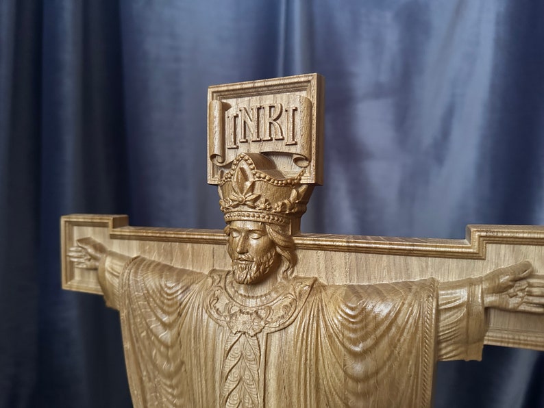 Może przedstawiać: Drewniany krucyfiks z wyrzeźbioną postacią Jezusa z wyciągniętymi ramionami. Postać nosi koronę i szatę. Na g&oacute;rze krzyża wyryto litery "INRI". Drewno ma naturalny, jasnobrązowy kolor.