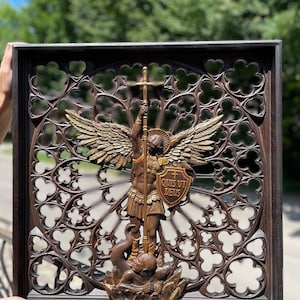 May include: A dark brown wooden wall plaque featuring a carved relief of Saint Michael the Archangel with wings, holding a spear and a shield inscribed with the Latin text QUIS UT DEUS, standing over a figure, set against a gothic trefoil pattern.