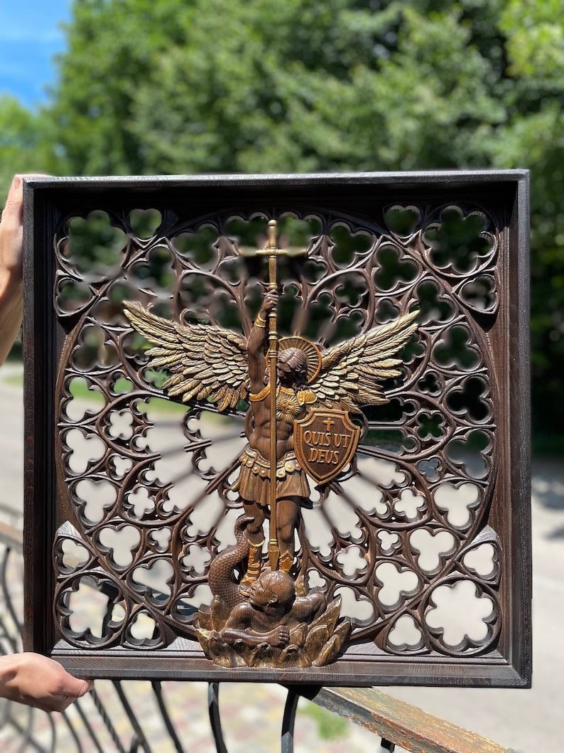 May include: A dark brown wooden wall plaque featuring a carved relief of Saint Michael the Archangel with wings, holding a spear and a shield inscribed with the Latin text QUIS UT DEUS, standing over a figure, set against a gothic trefoil pattern.