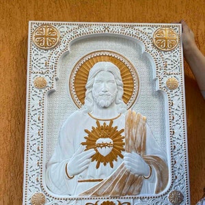 K&ouml;nnte beinhalten: Eine rechteckige Wandplatte in Wei&szlig; und Gold mit einem Relief des Heiligsten Herzens Jesu. Die zentrale Figur ist von einem verzierten Rahmen im Filigranstil umgeben, der an den Ecken goldene Akzente und einen strukturierten Hintergrund aufweist.