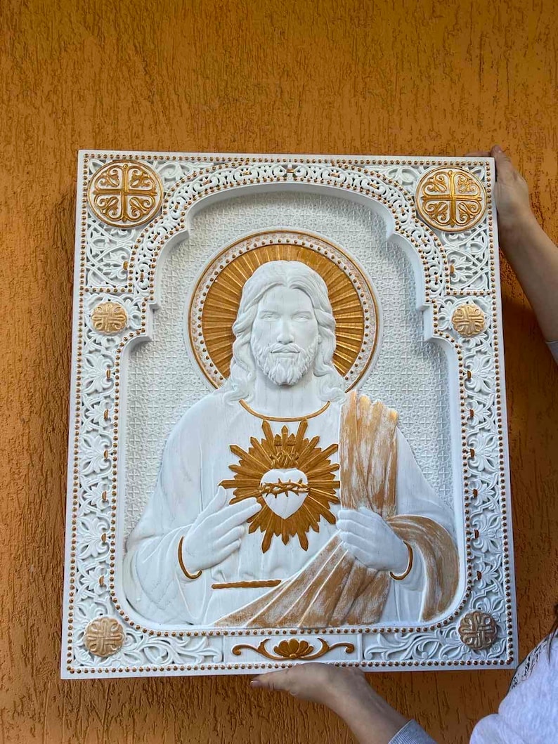 K&ouml;nnte beinhalten: Eine rechteckige Wandplatte in Wei&szlig; und Gold mit einem Relief des Heiligsten Herzens Jesu. Die zentrale Figur ist von einem verzierten Rahmen im Filigranstil umgeben, der an den Ecken goldene Akzente und einen strukturierten Hintergrund aufweist.