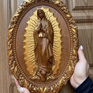 May include: A brown and gold oval-shaped religious wall plaque featuring a carved depiction of the Virgin Mary. The figure is surrounded by a gold halo and set against a wooden background. The plaque is framed with a detailed gold floral border.