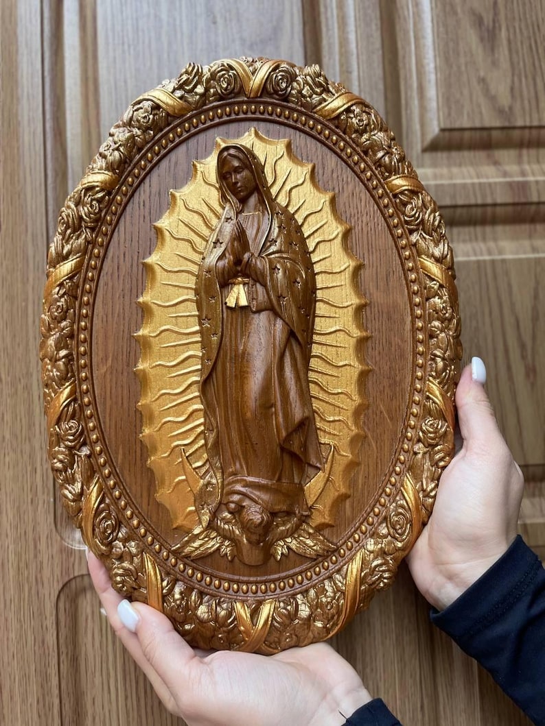 May include: A brown and gold oval-shaped religious wall plaque featuring a carved depiction of the Virgin Mary. The figure is surrounded by a gold halo and set against a wooden background. The plaque is framed with a detailed gold floral border.