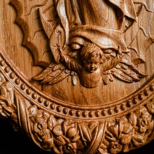 May include: A detailed, carved wooden relief featuring a religious scene. The artwork depicts an angel with outstretched wings, a figure with flowing robes, and ornate floral and beaded border. The wood has a warm, natural tone.