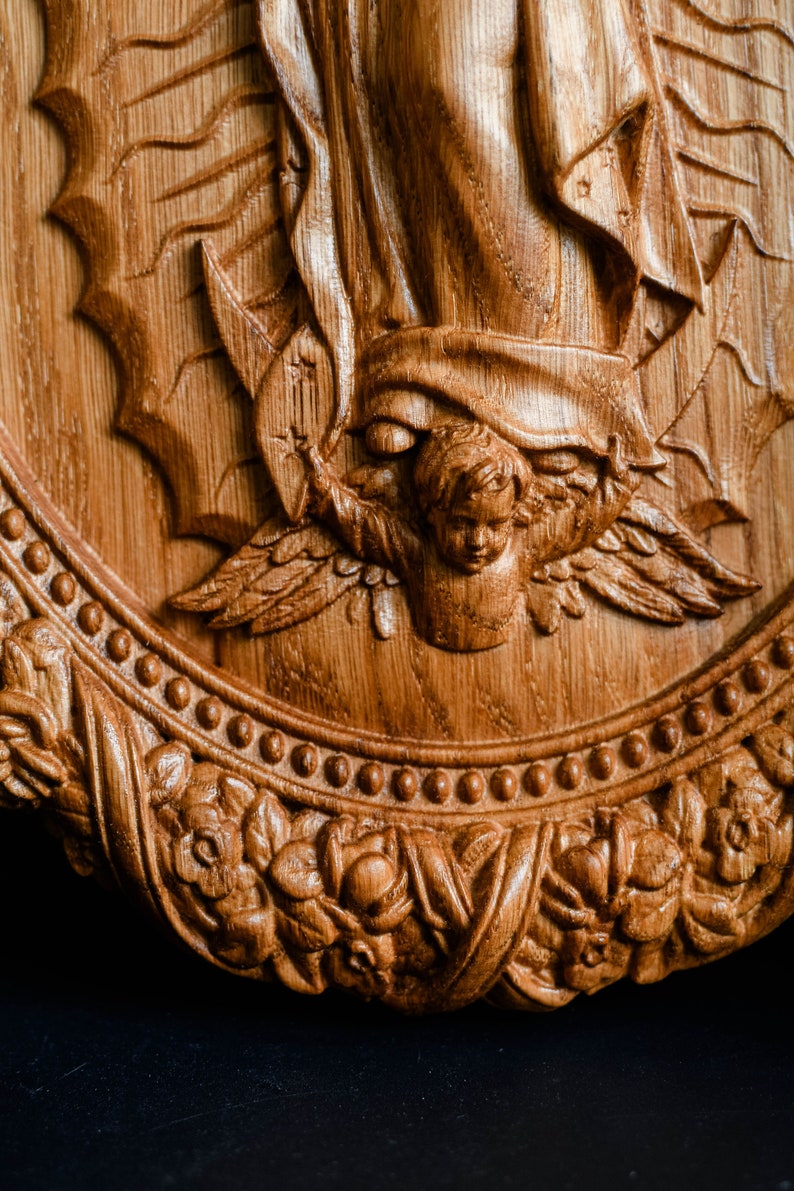 May include: A detailed, carved wooden relief featuring a religious scene. The artwork depicts an angel with outstretched wings, a figure with flowing robes, and ornate floral and beaded border. The wood has a warm, natural tone.