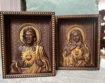 Puede incluir: Dos tallas de madera religiosas enmarcadas. La talla de la izquierda representa a Jesús con un corazón dorado y rayos de luz. La talla de la derecha representa a María con un corazón dorado y rayos de luz.