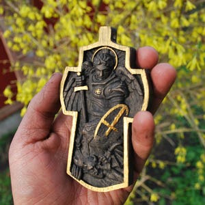 May include: A wooden cross with a carved relief of an angel holding a sword, with gold accents. The angel is wearing armor and has wings. The cross has a gold border and a gold letter "A".