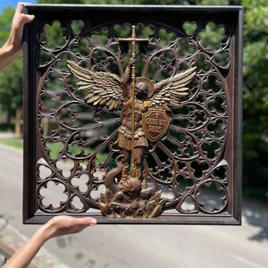 May include: A square dark brown wooden wall plaque featuring an intricate gothic-style openwork frame. In the center, a bronze-toned relief depicts an angel with wings holding a sword and a shield inscribed with the Latin text QUIS UT DEUS.