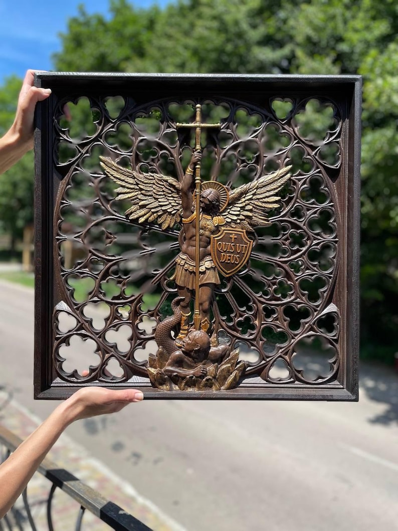 May include: A square dark brown wooden wall plaque featuring an intricate gothic-style openwork frame. In the center, a bronze-toned relief depicts an angel with wings holding a sword and a shield inscribed with the Latin text QUIS UT DEUS.