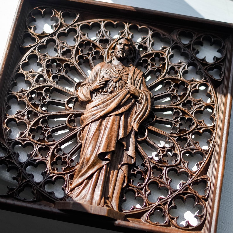 Puede incluir: Un adorno de pared de madera con una imagen tallada de una persona sosteniendo un coraz&oacute;n. El fondo es un dise&ntilde;o tallado que se asemeja a una ventana de vitral.