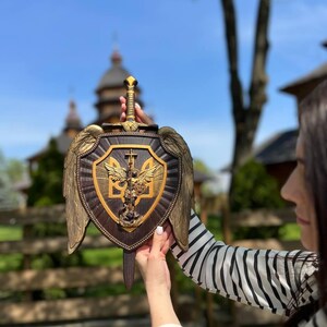 May include: A dark brown wooden shield wall plaque featuring a central gold-toned relief of Archangel Michael and the Ukrainian trident emblem. The shield is adorned with large gold-toned wings on both sides and a sword handle at the top.