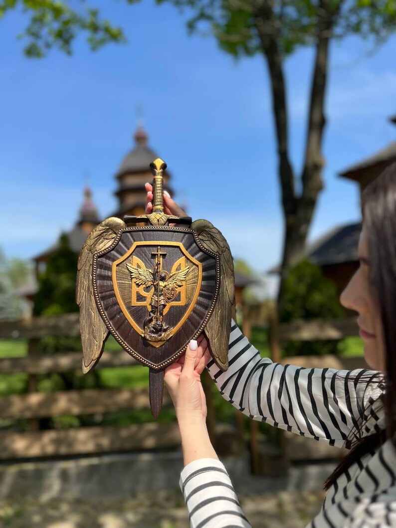 May include: A dark brown wooden shield wall plaque featuring a central gold-toned relief of Archangel Michael and the Ukrainian trident emblem. The shield is adorned with large gold-toned wings on both sides and a sword handle at the top.