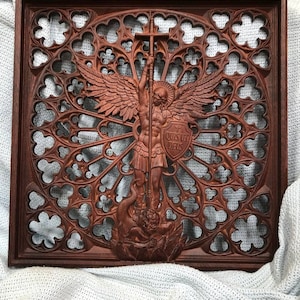May include: A dark brown wooden wall plaque featuring a detailed relief carving of an angel holding a cross and a shield with the Latin text 'QUIS UT DEUS'. The figure stands over a serpent, set against an intricate, openwork gothic rose window.