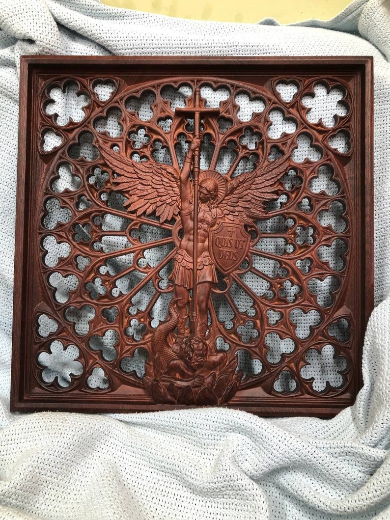 May include: A dark brown wooden wall plaque featuring a detailed relief carving of an angel holding a cross and a shield with the Latin text 'QUIS UT DEUS'. The figure stands over a serpent, set against an intricate, openwork gothic rose window.