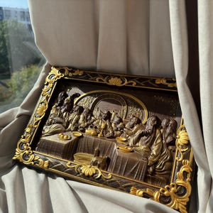 May include: A framed wood carving depicting the Last Supper. The carving is painted in brown and gold tones and features a detailed scene of Jesus and his disciples gathered around a table.