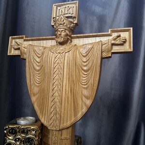 Może przedstawiać: Drewniana rzeźba krucyfiksu przedstawiająca ukoronowaną postać z wyciągniętymi ramionami, ubraną w szaty. Krzyż nosi napis "INRI". Rzeźba jest jasnobrązowa i spoczywa na dekoracyjnej podstawie.
